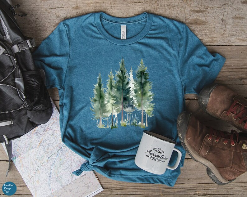 May include: Teal-colored t-shirt featuring a watercolor illustration of several evergreen trees. The shirt is paired with a white enamel mug that reads "And so the adventure begins." Hiking boots and a backpack are also visible.