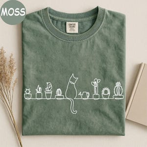 May include: A moss green crew neck t-shirt featuring a white continuous line drawing graphic. The design depicts a cat sitting among various potted cacti and a small watering can. The shirt has a Comfort Colors brand label at the inner neckline.