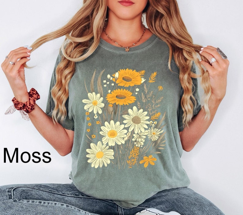 May include: Moss green t-shirt featuring a floral design with yellow sunflowers and white daisies. The graphic includes various wildflowers and the word "Moss" in the lower left corner. The shirt has short sleeves and a relaxed fit.