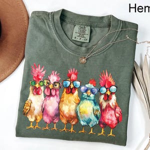 Puede incluir: Una camiseta verde con un gr&aacute;fico de cinco pollos con gafas de sol. Los pollos est&aacute;n pintados en un estilo acuarela y llevan gafas de sol de diferentes colores. La camiseta tiene el texto "COMFORT COLORS" y "S - P - CH" impreso en la parte delantera.