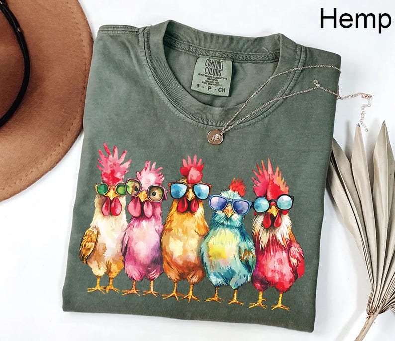 Puede incluir: Una camiseta verde con un gr&aacute;fico de cinco pollos con gafas de sol. Los pollos est&aacute;n pintados en un estilo acuarela y llevan gafas de sol de diferentes colores. La camiseta tiene el texto "COMFORT COLORS" y "S - P - CH" impreso en la parte delantera.