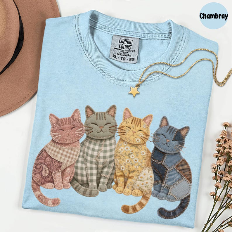 May include: Pale blue t-shirt featuring a design of four smiling cats in different patterns and colours. The shirt has a crew neck and a tag that says "Comfort Colors". A gold star pendant necklace is draped across the shirt.