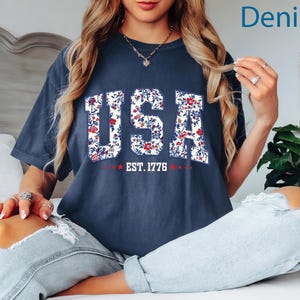 May include: A navy blue t-shirt with the word "USA" in a floral pattern. The text "EST. 1776" is below the word "USA" in white text with red stars. The shirt is a relaxed fit.