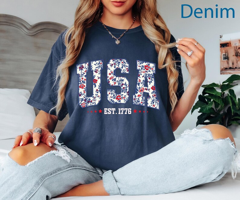 May include: A navy blue t-shirt with the word "USA" in a floral pattern. The text "EST. 1776" is below the word "USA" in white text with red stars. The shirt is a relaxed fit.