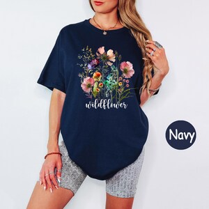 May include: Navy blue t-shirt featuring a watercolor floral design with pink, purple, and yellow flowers. The word "wildflower" is printed in white script below the flowers. The shirt has short sleeves and a relaxed fit.