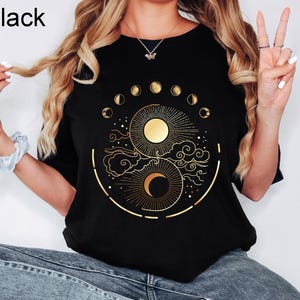 Pu&ograve; includere: T-shirt nera con un disegno dorato di sole e luna con fasi lunari. La maglietta ha un girocollo e maniche corte. La parola "Black" &egrave; stampata in alto a sinistra.