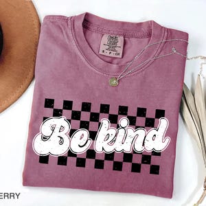 Pu&ograve; includere: Una t-shirt a maniche corte color bacca con una grafica a scacchi bianchi e neri e la scritta "Be kind" in un carattere bianco in stile retr&ograve; e dall'aspetto usurato. La maglietta presenta un girocollo e un'etichetta Comfort Colors.
