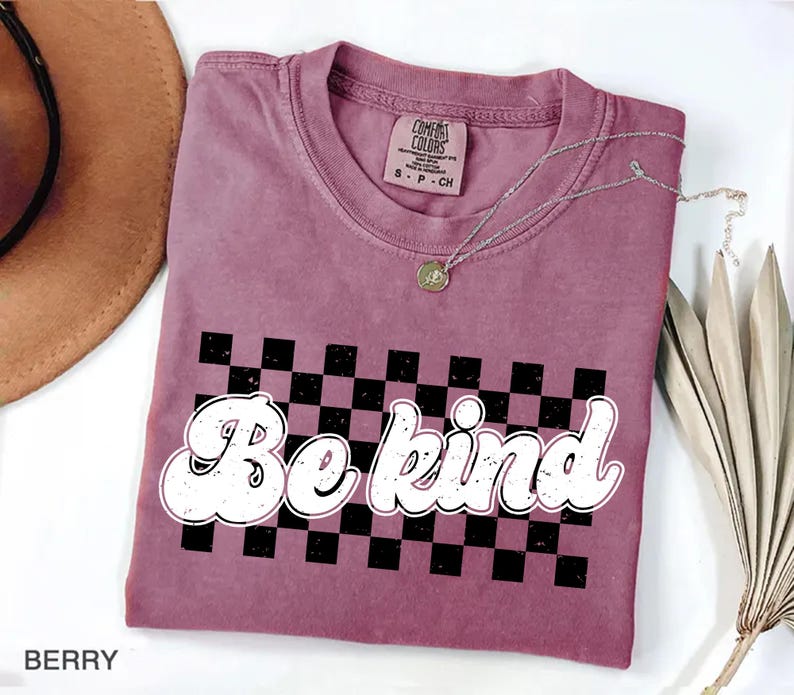 Pu&ograve; includere: Una t-shirt a maniche corte color bacca con una grafica a scacchi bianchi e neri e la scritta "Be kind" in un carattere bianco in stile retr&ograve; e dall'aspetto usurato. La maglietta presenta un girocollo e un'etichetta Comfort Colors.