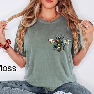 May include: Moss green t-shirt featuring a colorful floral bee design on the front. The bee has detailed wings and a body filled with flowers. The word "Moss" is printed in black on the bottom left of the image. The shirt is a casual style.