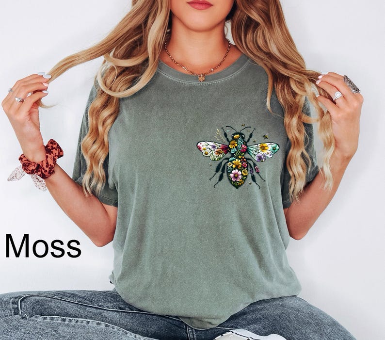 May include: Moss green t-shirt featuring a colorful floral bee design on the front. The bee has detailed wings and a body filled with flowers. The word "Moss" is printed in black on the bottom left of the image. The shirt is a casual style.