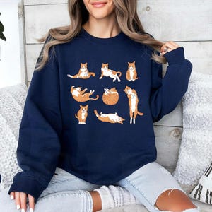 May include: A navy blue crewneck sweatshirt featuring a graphic print of eight orange and white tabby cats in various lounging and stretching poses. The cats are arranged in a grid-like pattern across the center of the chest area.