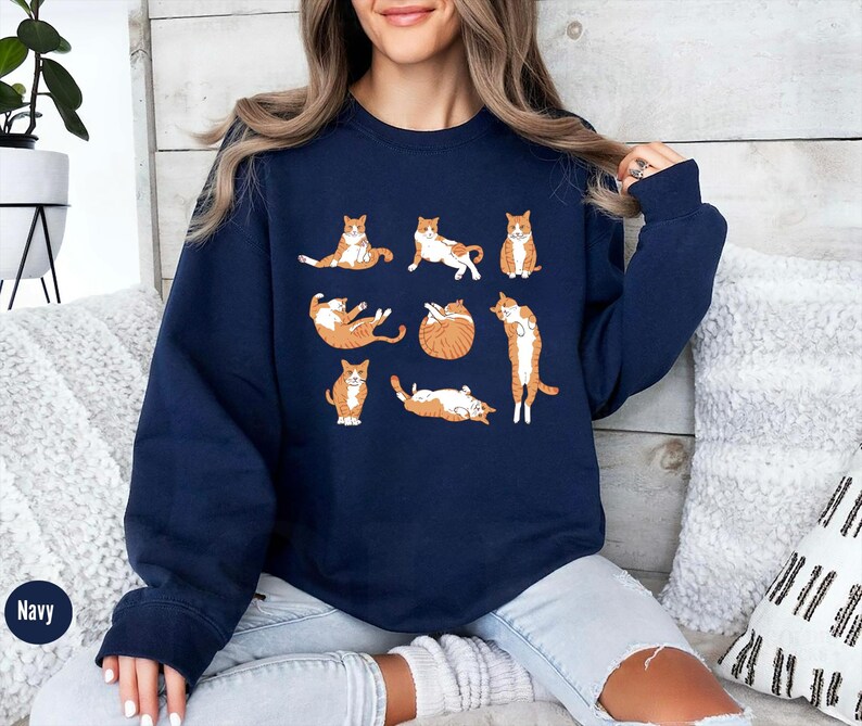 May include: A navy blue crewneck sweatshirt featuring a graphic print of eight orange and white tabby cats in various lounging and stretching poses. The cats are arranged in a grid-like pattern across the center of the chest area.