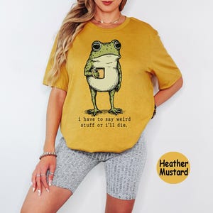 May include: A heather mustard short-sleeve t-shirt featuring a green frog illustration holding a small mug. Below the frog, the text reads: i have to say weird stuff or i'll die. The shirt is paired with grey ribbed shorts.