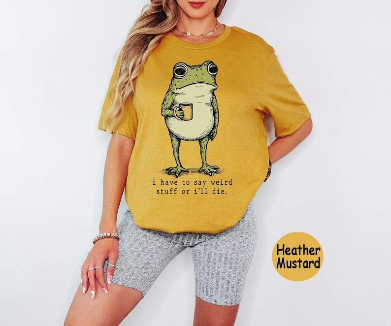May include: A heather mustard short-sleeve t-shirt featuring a green frog illustration holding a small mug. Below the frog, the text reads: i have to say weird stuff or i'll die. The shirt is paired with grey ribbed shorts.