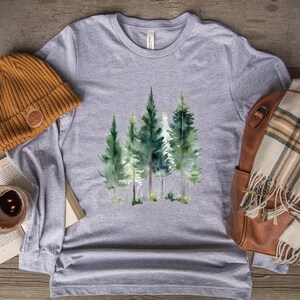 May include: A heather gray long-sleeve shirt featuring a watercolor design of evergreen trees. A brown beanie, a book, a candle, a brown leather bag, and a plaid scarf are also visible. The shirt has the text "Athletic Heather".