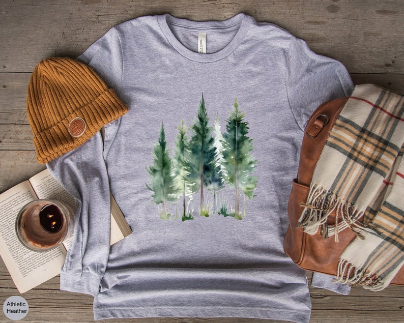 May include: A heather gray long-sleeve shirt featuring a watercolor design of evergreen trees. A brown beanie, a book, a candle, a brown leather bag, and a plaid scarf are also visible. The shirt has the text "Athletic Heather".