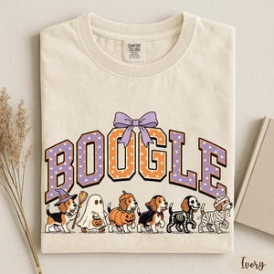 K&ouml;nnte beinhalten: Ein elfenbeinfarbenes T-Shirt mit dem Schriftzug BOOGLE in gro&szlig;en orangefarbenen und violetten gepunkteten Buchstaben, gekr&ouml;nt von einer violetten Schleife. Darunter sind sechs Beagle-Hunde in Halloween-Kost&uuml;men als Hexe, Geist, K&uuml;rbis, Vampir, Skelett und Mumie abgebildet.