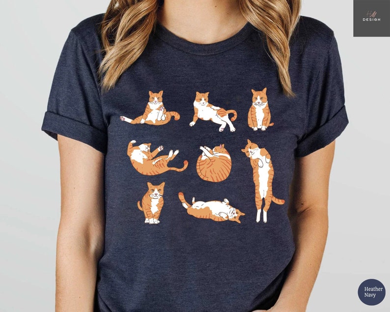 May include: Heather navy t-shirt featuring a playful illustration of orange and white cats in various poses. The design includes cats sitting, stretching, and curled up. The shirt has a classic crew neck and short sleeves.