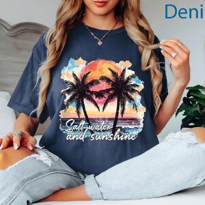 May include: Denim blue t-shirt with a colorful graphic of a sunset, palm trees, and the text "Salt water and sunshine." The shirt is a relaxed fit.