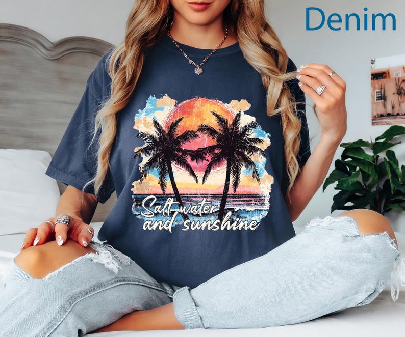 May include: Denim blue t-shirt with a colorful graphic of a sunset, palm trees, and the text "Salt water and sunshine." The shirt is a relaxed fit.