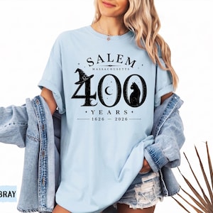 Puede incluir: Una camiseta azul claro con un estampado gr&aacute;fico negro que dice SALEM MASSACHUSETTS 400 YEARS 1626-2026. El dise&ntilde;o incluye un sombrero de bruja sobre el n&uacute;mero cuatro, una luna creciente en el cero y la silueta de un gato negro en el cero.
