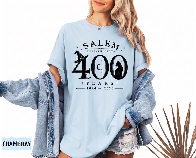 Puede incluir: Una camiseta azul claro con un estampado gr&aacute;fico negro que dice SALEM MASSACHUSETTS 400 YEARS 1626-2026. El dise&ntilde;o incluye un sombrero de bruja sobre el n&uacute;mero cuatro, una luna creciente en el cero y la silueta de un gato negro en el cero.