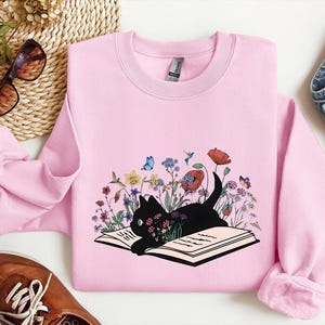 May include: A light pink sweatshirt featuring a black cat lying on an open book, surrounded by colorful flowers and butterflies. The graphic design includes red poppies, blue butterflies, and various wildflowers. The sweatshirt has a crew neck and long sleeves.