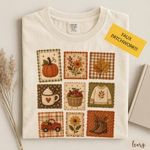 May include: An ivory t-shirt featuring a grid of nine square patches with autumn-themed illustrations including a pumpkin, flowers, a leaf, a mug with a heart, a basket of apples, a sweater, a red truck, a sunflower, and boots.