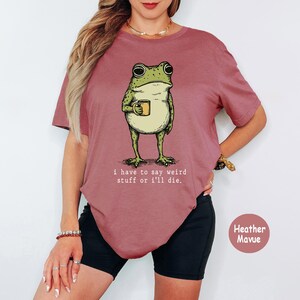 May include: A heather mauve t-shirt featuring a graphic of a green frog holding a small yellow mug. Below the frog, the text reads: i have to say weird stuff or i'll die. The shirt has short sleeves and a crew neckline, presented in a solid colour.