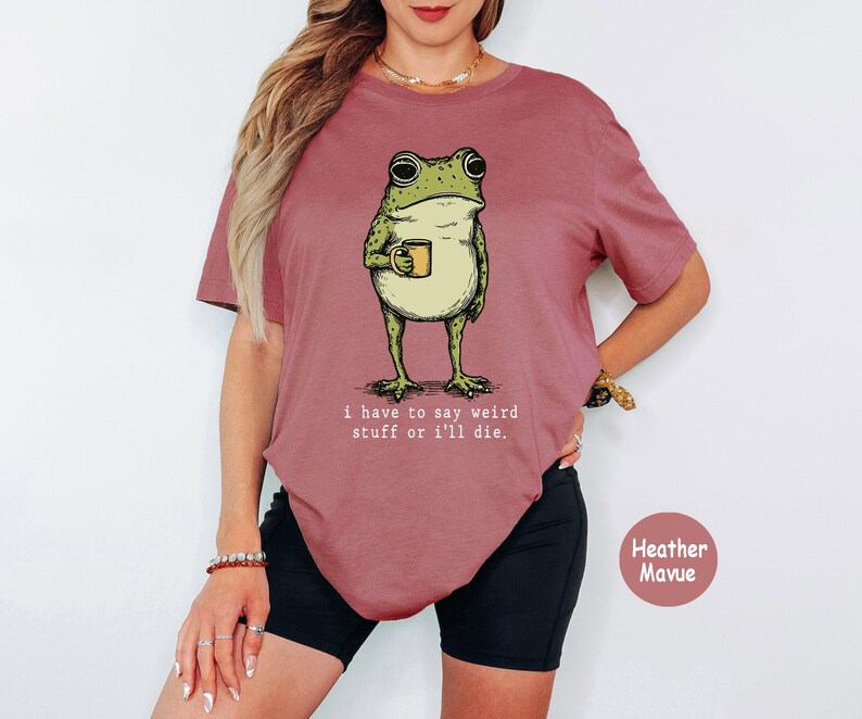May include: A heather mauve t-shirt featuring a graphic of a green frog holding a small yellow mug. Below the frog, the text reads: i have to say weird stuff or i'll die. The shirt has short sleeves and a crew neckline, presented in a solid colour.