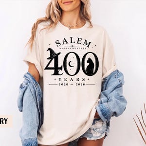 Puede incluir: Una camiseta de color marfil con texto y gr&aacute;ficos en negro que dicen SALEM MASSACHUSETTS 400 YEARS 1626-2026. El dise&ntilde;o incluye un sombrero de bruja sobre el n&uacute;mero 4, una luna creciente en el cero y la silueta de un gato negro en el &uacute;ltimo cero.