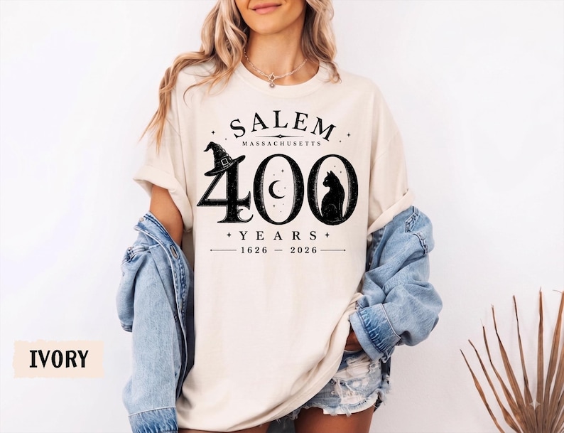 Puede incluir: Una camiseta de color marfil con texto y gr&aacute;ficos en negro que dicen SALEM MASSACHUSETTS 400 YEARS 1626-2026. El dise&ntilde;o incluye un sombrero de bruja sobre el n&uacute;mero 4, una luna creciente en el cero y la silueta de un gato negro en el &uacute;ltimo cero.