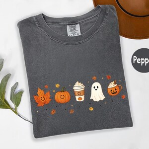 K&ouml;nnte beinhalten: Ein anthrazitfarbenes T-Shirt mit einem Grafikdruck, der ein l&auml;chelndes orangefarbenes Ahornblatt, einen l&auml;chelnden K&uuml;rbis, einen Kaffeebecher, ein wei&szlig;es Gespenst und eine K&uuml;rbistasse zeigt, umgeben von kleinen Herbstbl&auml;ttern und Sternen.