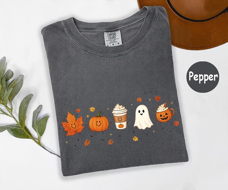 K&ouml;nnte beinhalten: Ein anthrazitfarbenes T-Shirt mit einem Grafikdruck, der ein l&auml;chelndes orangefarbenes Ahornblatt, einen l&auml;chelnden K&uuml;rbis, einen Kaffeebecher, ein wei&szlig;es Gespenst und eine K&uuml;rbistasse zeigt, umgeben von kleinen Herbstbl&auml;ttern und Sternen.