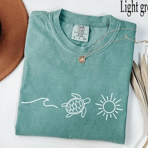 May include: A light green short-sleeved t-shirt featuring a white graphic print of a wave, a sea turtle, and a sun. The shirt has a crew neckline and a Comfort Colors brand label on the inside collar. The text Light green is visible at the top.