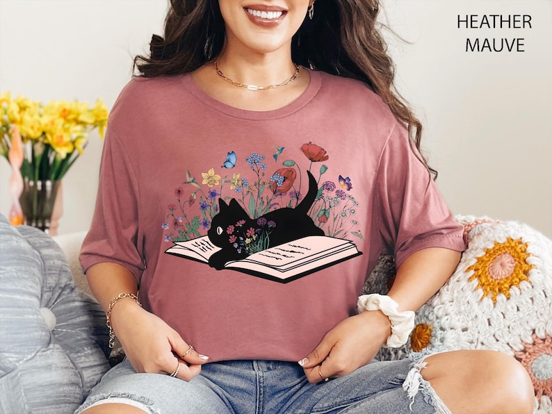 May include: Heather mauve t-shirt featuring a black cat lying on an open book, surrounded by colorful flowers and butterflies. The shirt has short sleeves and a round neckline. The text "HEATHER MAUVE" is printed on the upper right.