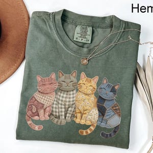 May include: Hemp green t-shirt with four smiling cat illustrations in patchwork patterns. The shirt has a crew neck and short sleeves. A silver necklace with a small pendant is draped across the shirt.