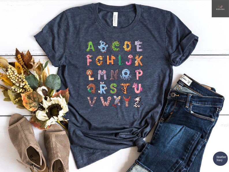 May include: Heather navy t-shirt featuring the alphabet, with each letter designed as a different animal. The letters are in various colors, including green, orange, and pink. The shirt is paired with blue jeans.