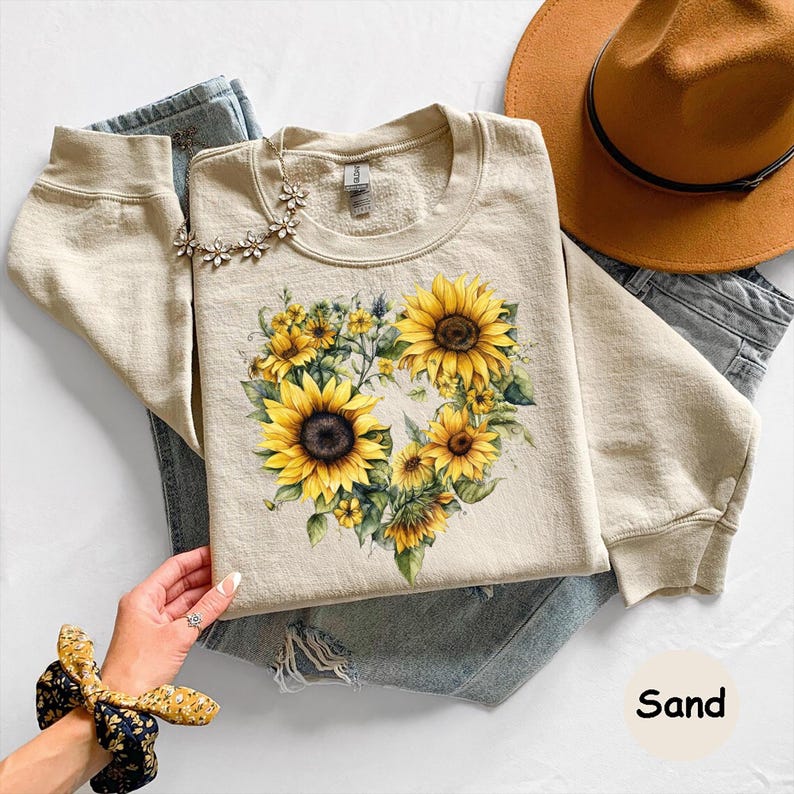 Pode incluir: Uma camisola de cor areia com um design de girass&oacute;is em forma de cora&ccedil;&atilde;o. O design inclui girass&oacute;is amarelos com centros castanhos e folhas verdes. A camisola &eacute; combinada com jeans azuis e um chap&eacute;u castanho.
