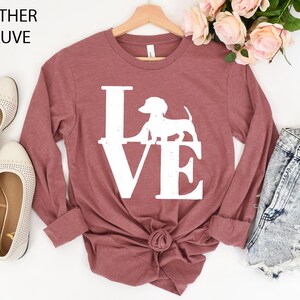 May include: Heather mauve long-sleeve shirt with a white "LOVE" graphic featuring a dachshund silhouette. The shirt is styled with a knot at the waist and is displayed with denim shorts and flats. The text "Heather Mauve" is at the top.
