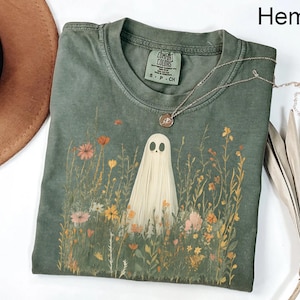 May include: A green Comfort Colors t-shirt featuring a ghost in a field of wildflowers. The ghost is white, and the flowers are in shades of pink, yellow, and orange. A silver necklace with a pendant is on the shirt. The word "Hemp" is in the top right corner.