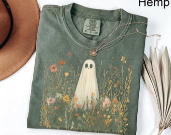 May include: A sage green t-shirt with a crew neckline and a graphic print of a white ghost standing in a field of colourful wildflowers. The shirt features a Comfort Colors brand label at the inner neck with the text S-P-CH and the word Hemp in the corner.