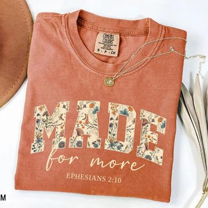 May include: A terracotta-colored t-shirt with the words "MADE for more" in a floral design. The text "Ephesians 2:10" is below the main text. A gold necklace with a pendant is on the shirt.