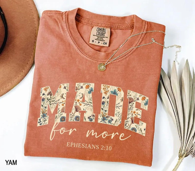 May include: A terracotta-colored t-shirt with the words "MADE for more" in a floral design. The text "Ephesians 2:10" is below the main text. A gold necklace with a pendant is on the shirt.