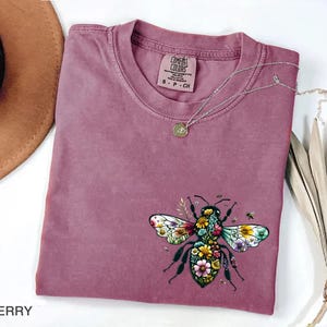 May include: Berry-colored t-shirt with a floral bee graphic. The bee design features colorful flowers and leaves, with a small Comfort Colors tag at the neckline. A silver necklace with a pendant is draped across the shirt.