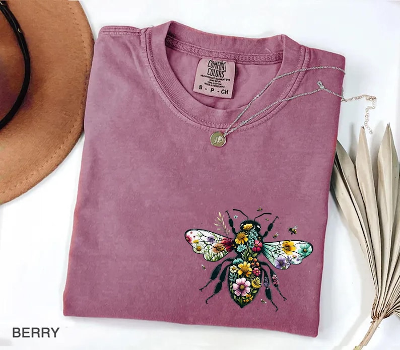 May include: Berry-colored t-shirt with a floral bee graphic. The bee design features colorful flowers and leaves, with a small Comfort Colors tag at the neckline. A silver necklace with a pendant is draped across the shirt.