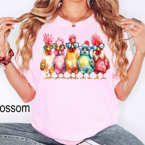 Puede incluir: Una camiseta rosa con un gr&aacute;fico de cinco gallos coloridos que llevan gafas de sol. Los gallos est&aacute;n dispuestos en fila y miran hacia adelante. Los gallos son de diferentes colores, incluyendo rojo, naranja, amarillo, verde y azul.