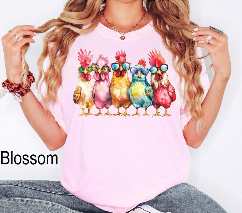 Puede incluir: Una camiseta rosa con un gr&aacute;fico de cinco gallos coloridos que llevan gafas de sol. Los gallos est&aacute;n dispuestos en fila y miran hacia adelante. Los gallos son de diferentes colores, incluyendo rojo, naranja, amarillo, verde y azul.