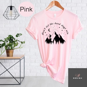 Pu&ograve; includere: T-shirt rosa chiaro con la scritta "Don't let the hard days win" sopra una grafica di montagna. Il design include una luna crescente, stelle e raggi di sole. La maglietta &egrave; appesa a una gruccia di legno.