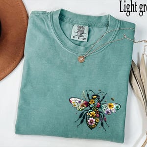 May include: Light green t-shirt with a floral bee graphic. The bee design features colorful flowers and detailed wings. A silver necklace with a small pendant rests on the shirt. The shirt is made of a soft material.